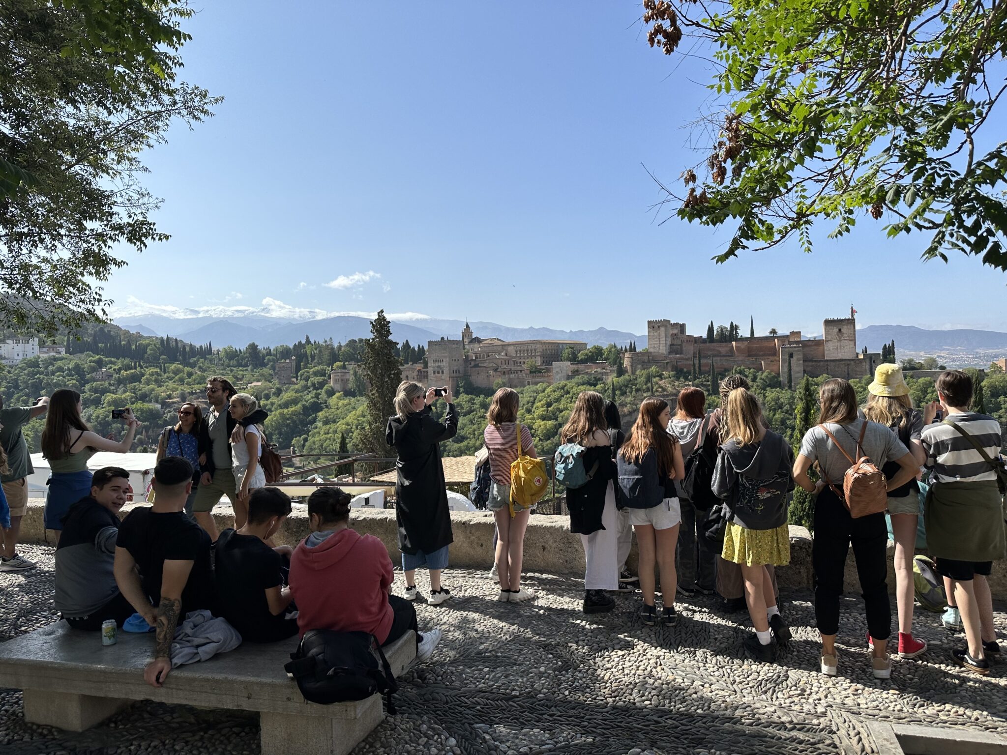Tourists enjoying the scenic view of the Alhambra with mountainous landscape in the background under a clear blue sky.