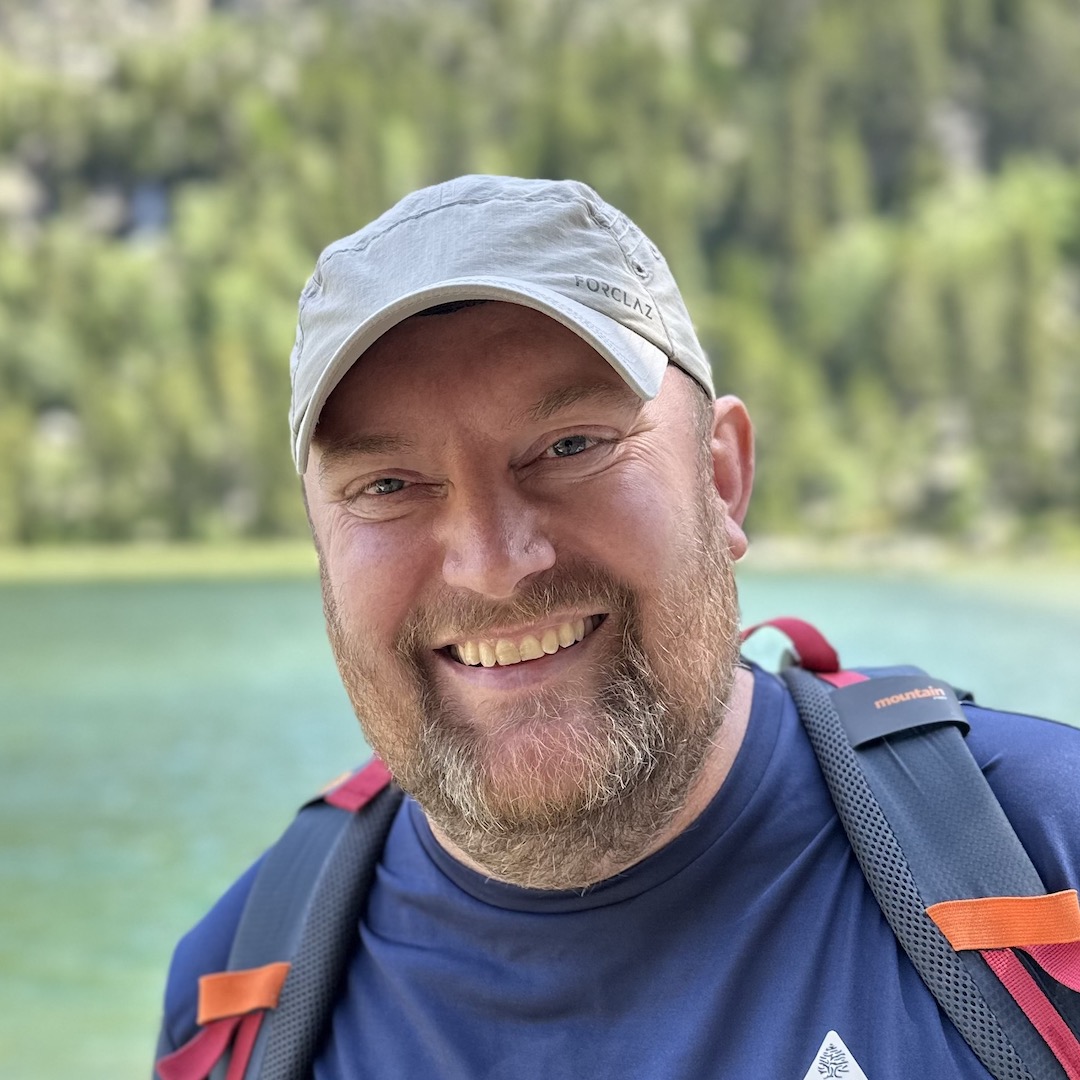 Smiling man in a gray cap and backpack outdoors near a lake with trees in the background.